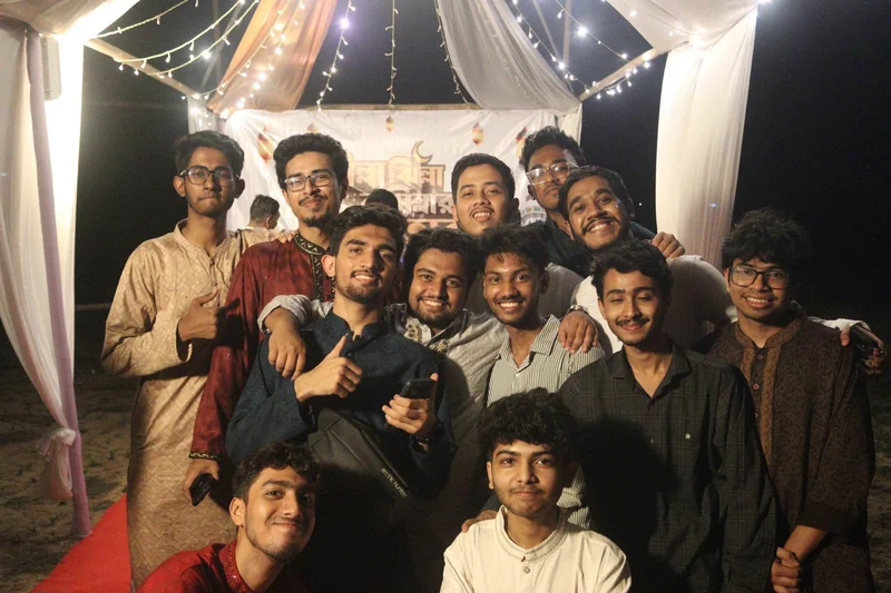 A cheerful group of young men in panjabis crowd together for a selfie-style photo under the lit iftar tent at night.