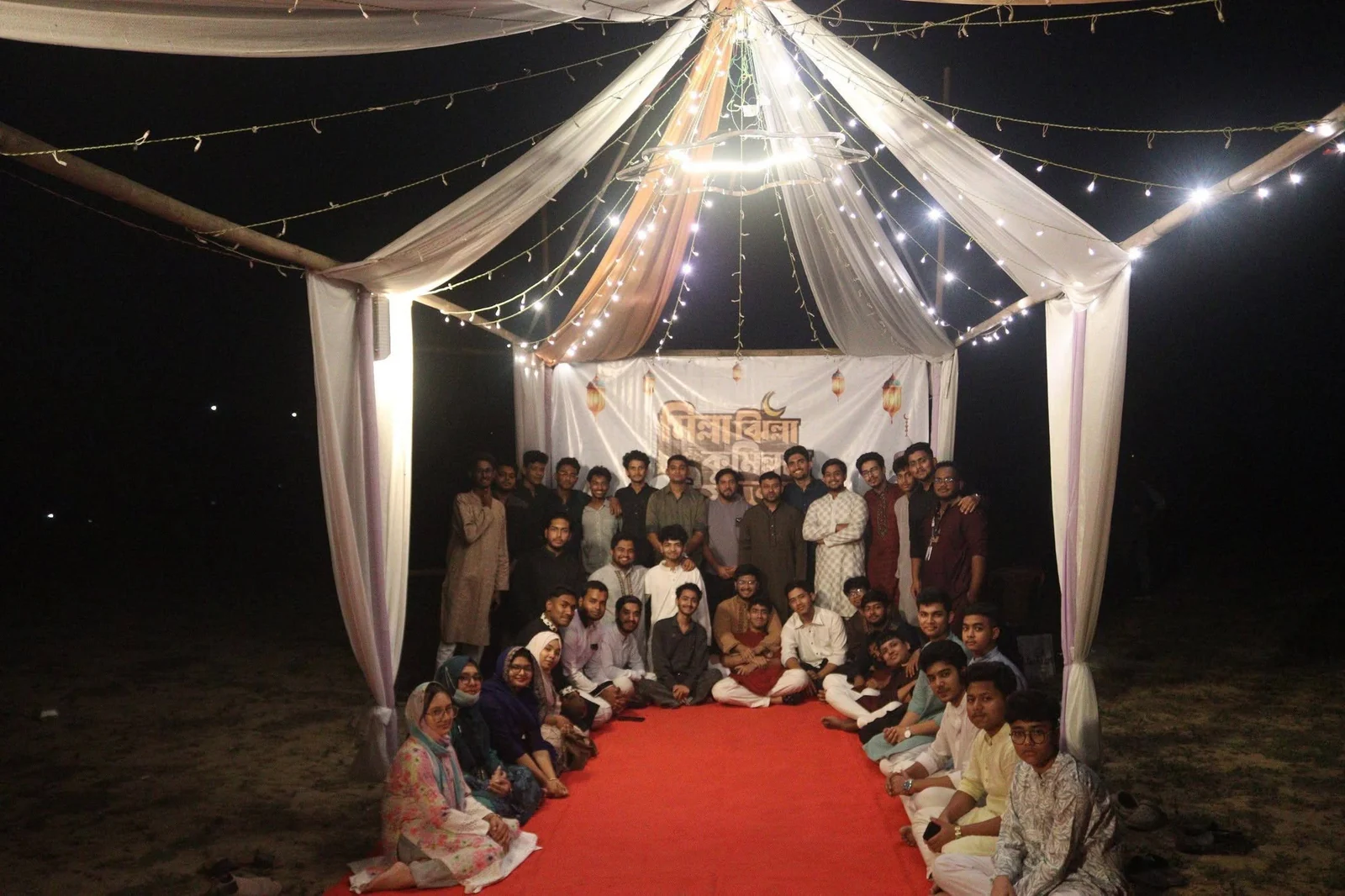 Around fifty young volunteers from TECHious and partner youth groups pose together on a red carpet under a white tent lit with string lights at night, Gomtir Par, Cumilla.