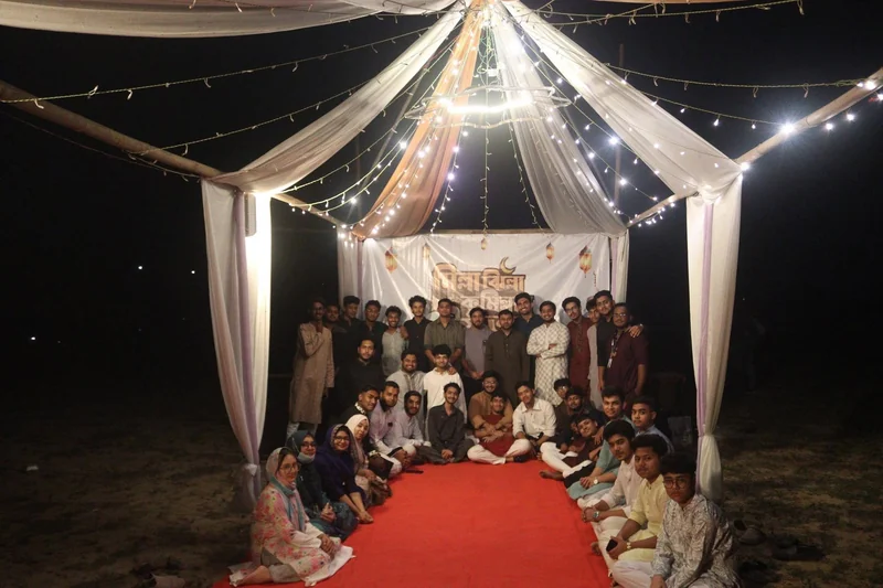 Around fifty young volunteers from TECHious and partner youth groups pose together on a red carpet under a white tent lit with string lights at night, Gomtir Par, Cumilla.