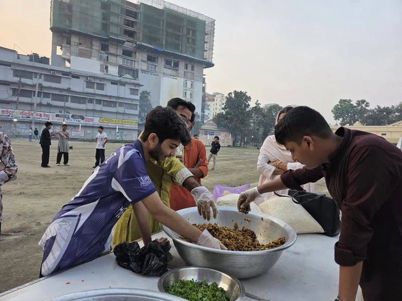 Young volunteers in gloves mix chickpea chola in large steel bowls on an outdoor table at sunset, with the Town Hall field behind.