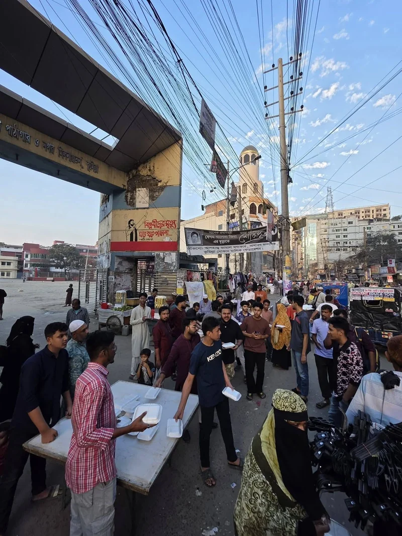 Volunteers set up an iftar distribution table under the arched gate of Cumilla Town Hall as people gather beneath a blue sky.