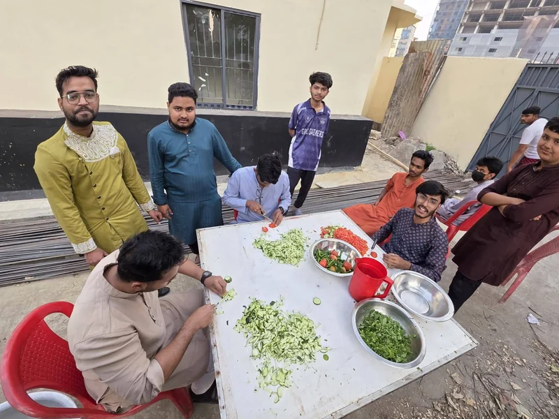 A team of young men chops cucumber and herbs at a long white table outdoors, preparing iftar salad.