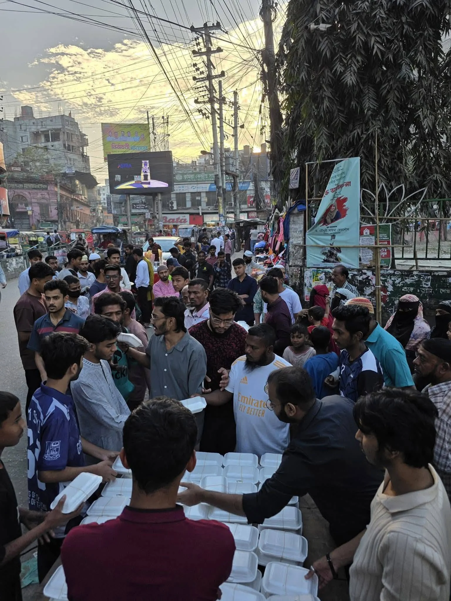 Volunteers hand out boxed iftar meals to a dense crowd on the street outside Cumilla Town Hall at dusk, with shops and power lines behind.