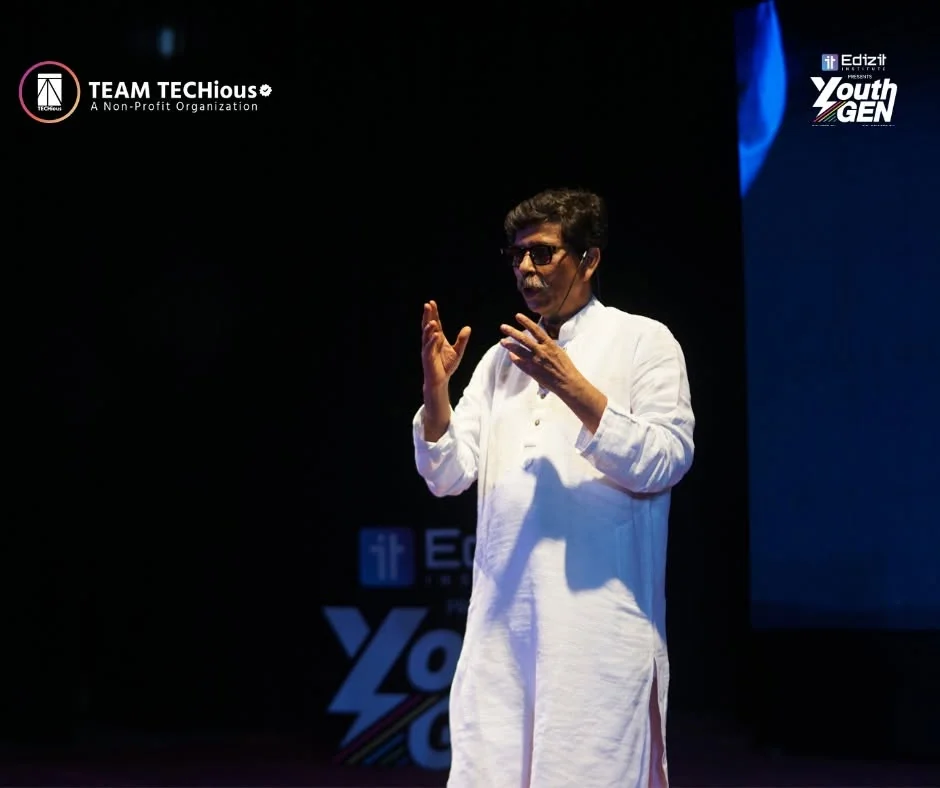 Hajee Aminur Rashid Yasin, in a white panjabi and glasses, gestures with both hands as he speaks on the dark YouthGEN 2025 stage.
