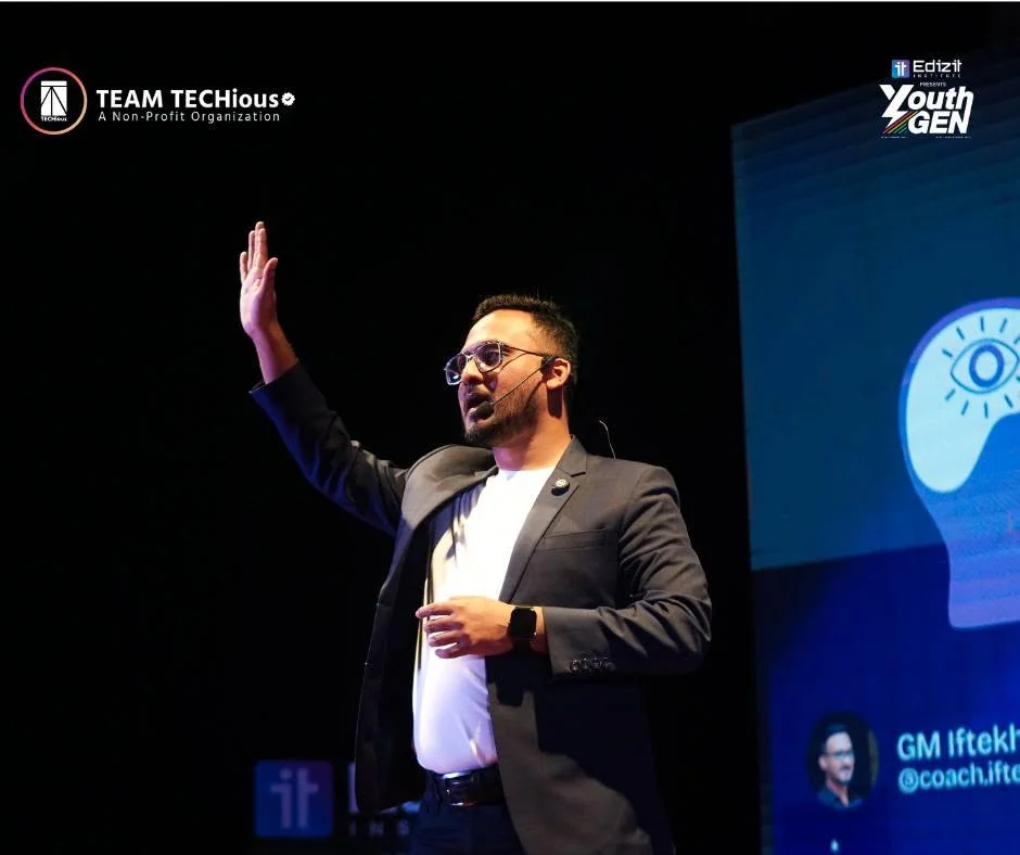 GM Iftekhar Iftee, in a grey blazer and white T-shirt, raises one hand as he speaks on the YouthGEN 2025 stage beside a screen with a lightbulb graphic.