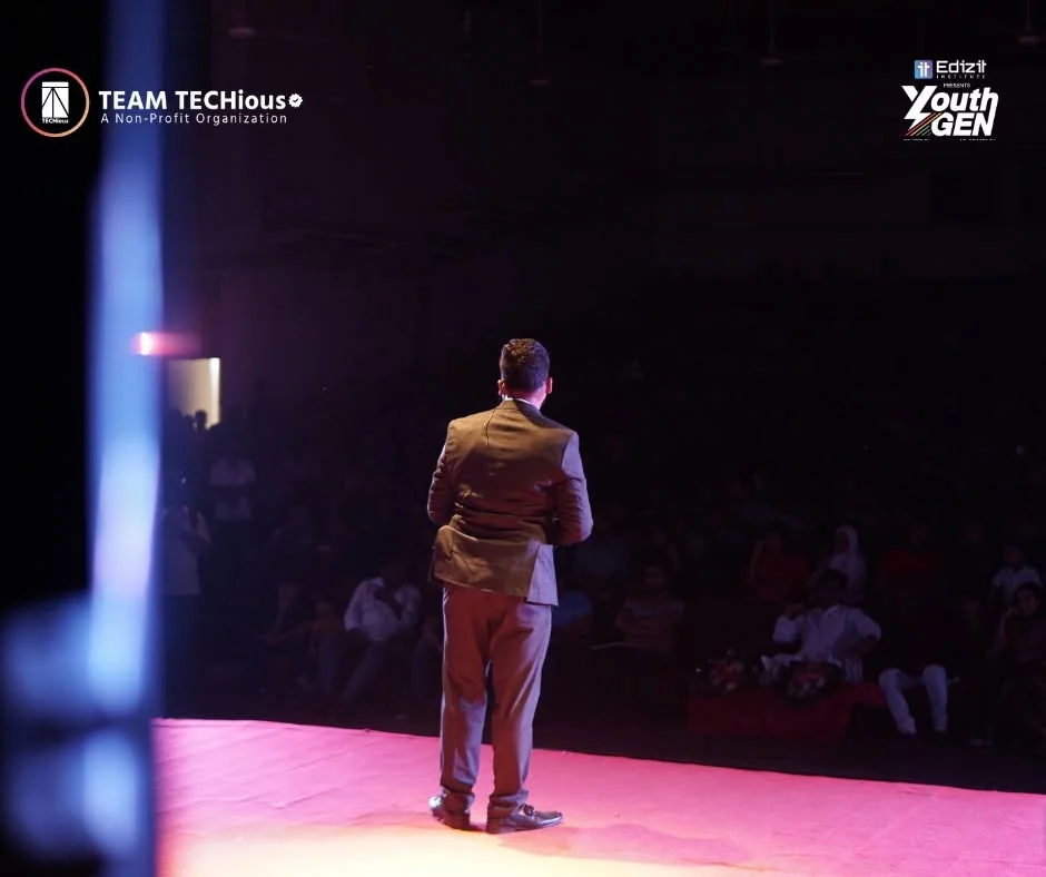 A speaker stands on a pink-lit stage seen from behind, facing a full auditorium at Shilpakala Academy.
