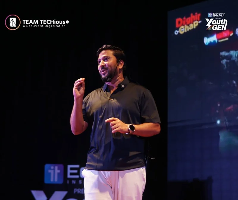 Kausar Akash, in a black polo shirt, speaks on the YouthGEN 2025 stage with the Dighir Chap logo on the screen behind him.