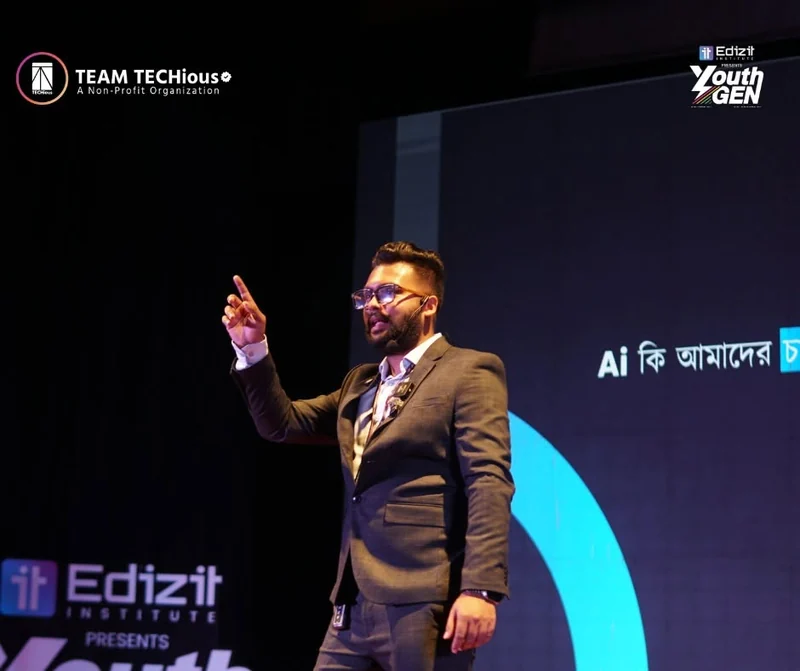 Mahi Rahman, in a dark suit, points upward on stage next to a screen with a Bangla question about AI and the Edizit Institute logo.