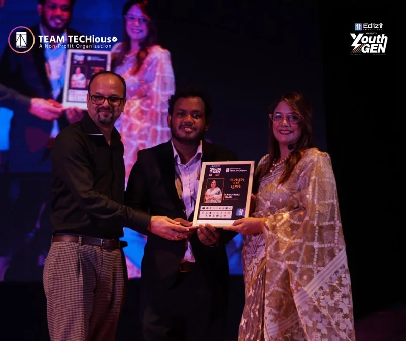 Farah Naz Islam Shila, in a cream sari, receives a framed Token of Love from two men on the YouthGEN 2025 stage.