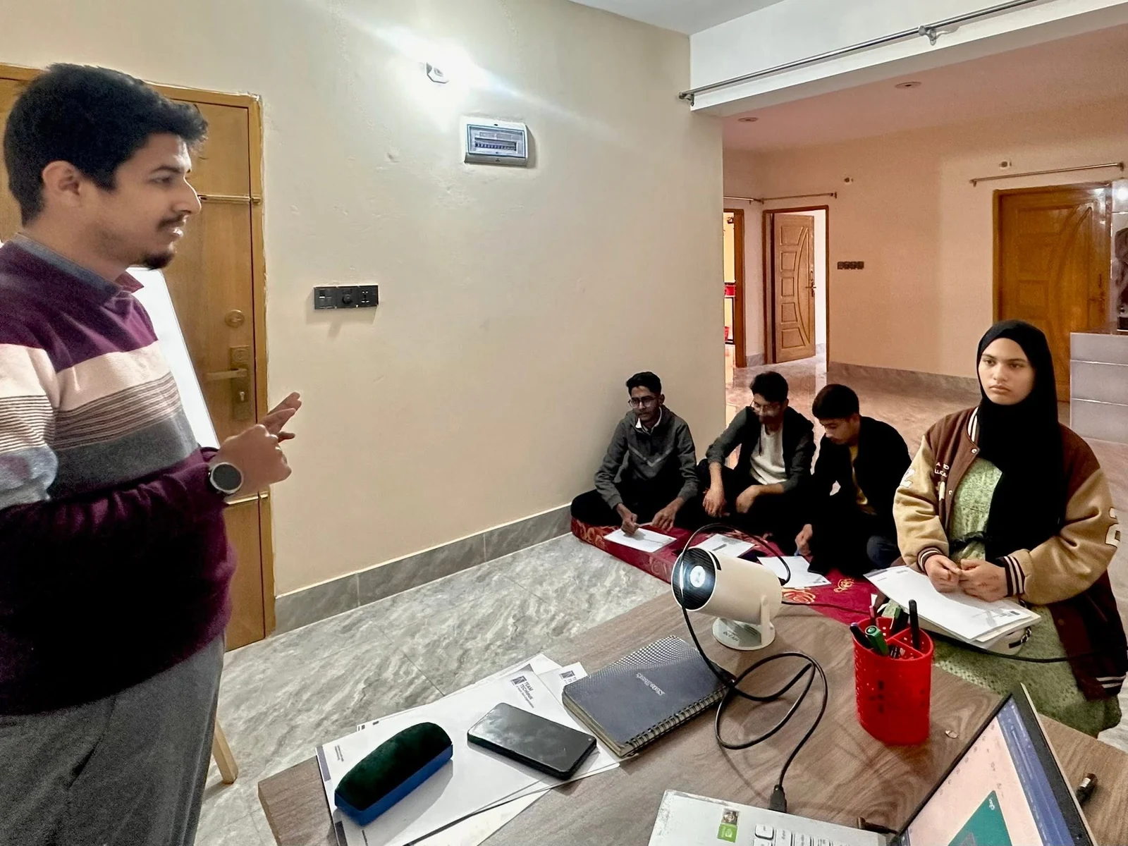 A trainer stands and speaks to a small group of learners seated on a floor mat with laptops and a projector in a bright room.
