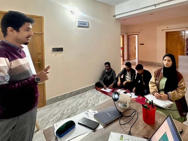 A trainer stands and speaks to a small group of learners seated on a floor mat with laptops and a projector in a bright room.