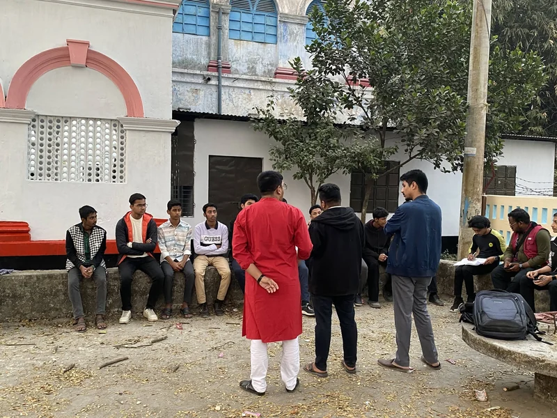 TECHious members sit on a low stone wall in a courtyard while a leader in a red panjabi addresses them during an outdoor meeting.