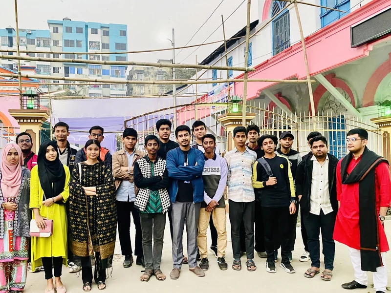 About twenty TECHious members, young men and women in casual and traditional clothes, stand in a line in front of a pink-and-white building with bamboo scaffolding in Cumilla.
