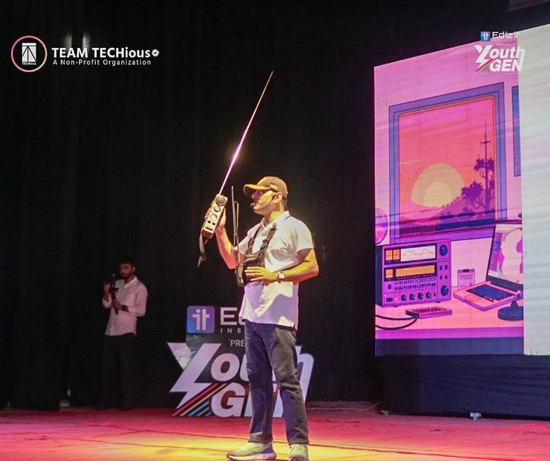 A man holds up a tall antenna on the YouthGEN 2025 stage during an amateur radio demonstration, with a retro radio graphic on the screen behind.
