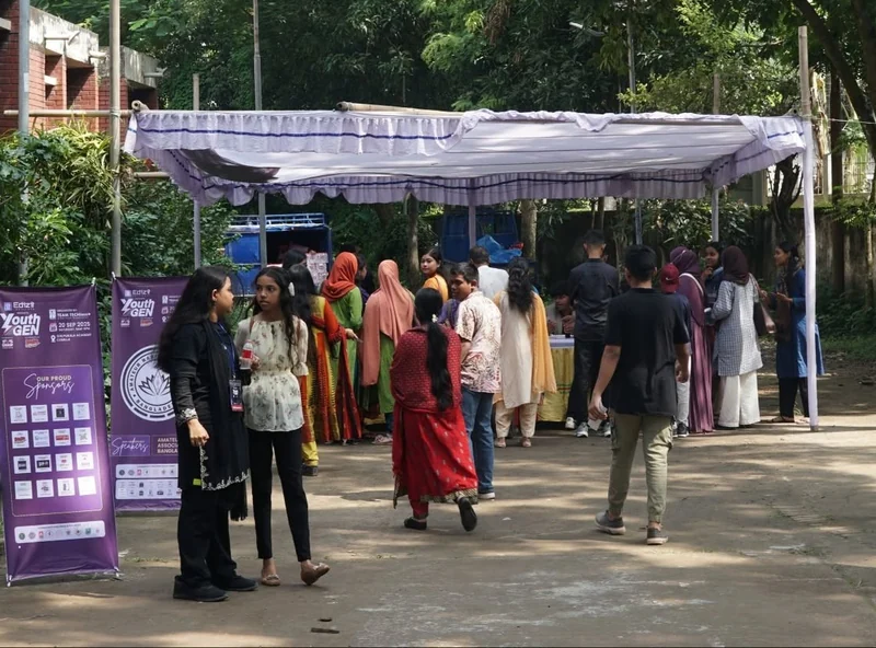 Visitors walk along a tree-lined path toward a white registration tent at Shilpakala Academy, Cumilla, past purple YouthGEN 2025 banners.