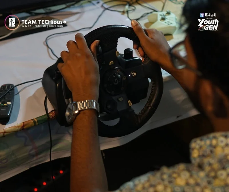 Close-up of hands on a racing-simulator steering wheel in the YouthGEN 2025 Experience Zone, with a monitor showing a driving game.