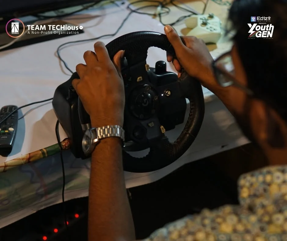 Close-up of hands on a racing-simulator steering wheel in the YouthGEN 2025 Experience Zone, with a monitor showing a driving game.