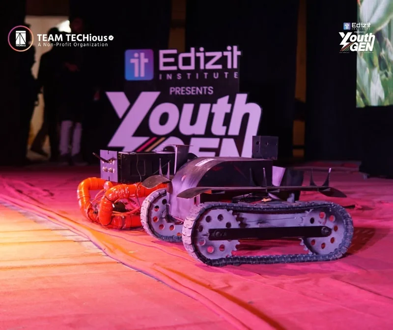 A tracked rover robot from BRAC University team Mongol-Tori sits on a pink-lit stage in front of the Edizit presents YouthGEN screen.