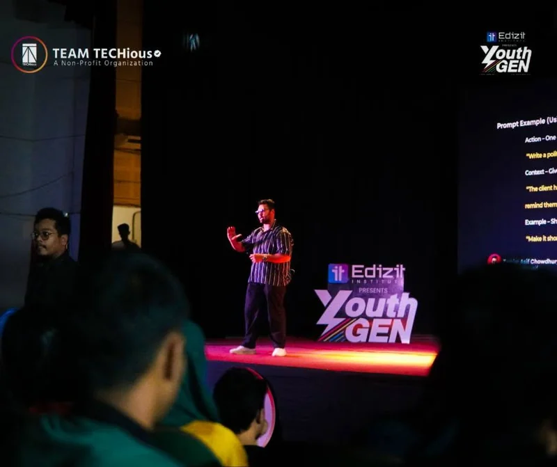 A speaker gestures on a dark stage in front of the YouthGEN 2025 logo and a screen about AI, with audience silhouettes below.