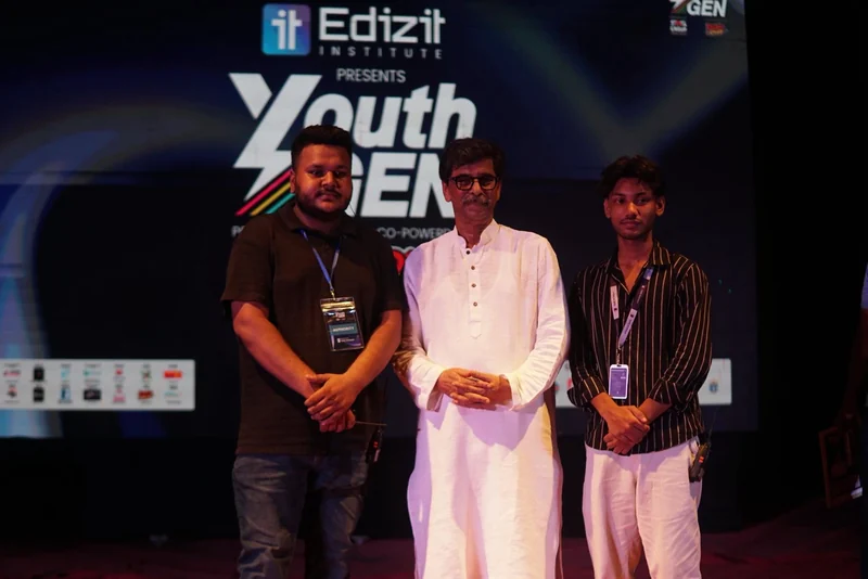 Three men stand on the dark YouthGEN 2025 stage in front of a large screen showing the Edizit presents YouthGEN logo; the guest in the centre wears a white panjabi.