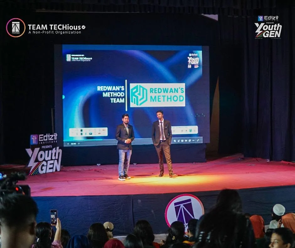 Wide view of the YouthGEN 2025 stage: two speakers stand under a spotlight in front of a large blue LED screen, with the audience in the foreground.