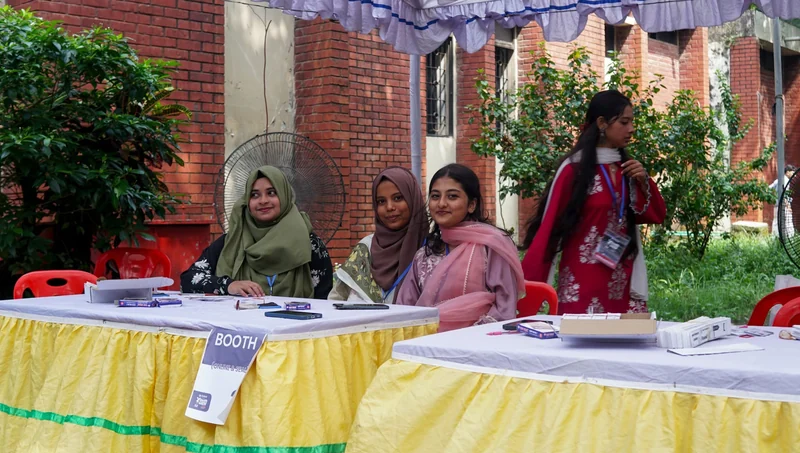 Young women volunteers in lanyards sit at a yellow-skirted ticket desk under a tent, checking in guests for YouthGEN 2025.